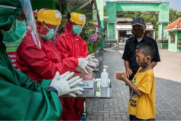 Profissionais de saúde com roupas de proteção ajudam um menino a usar álcool em gel em um posto de saúde comunitário.